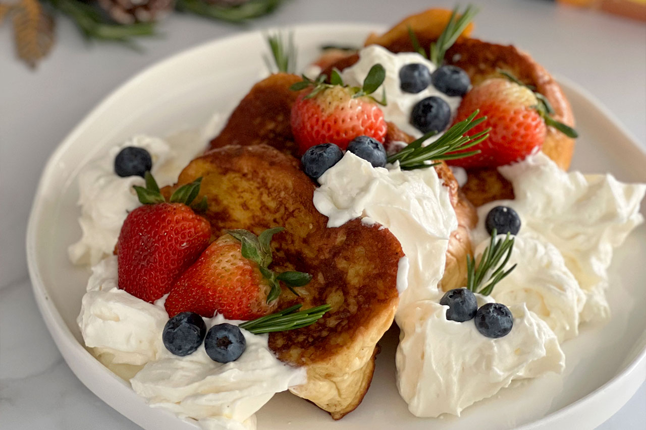 Tres rebanadas de pan francés doradas servidas en un plato blanco, cubiertas con crema batida, fresas frescas, arándanos y pequeñas ramas de romero como decoración