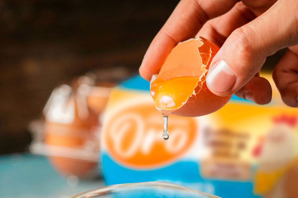Una mano sosteniendo la cáscara de un huevo marrón recién roto, dejando caer la yema dorada y parte de la clara hacia un recipiente
