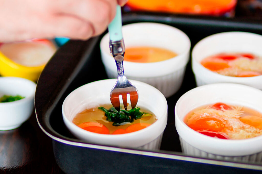 Una mano usa un tenedor para añadir espinacas a un recipiente con huevos crudos en un baño maría