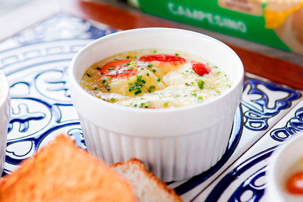 Primer plano de un ramequín de cerámica blanca con huevos al horno (shirred eggs), acompañados de tomates cherry, hierbas frescas picadas y queso derretido.