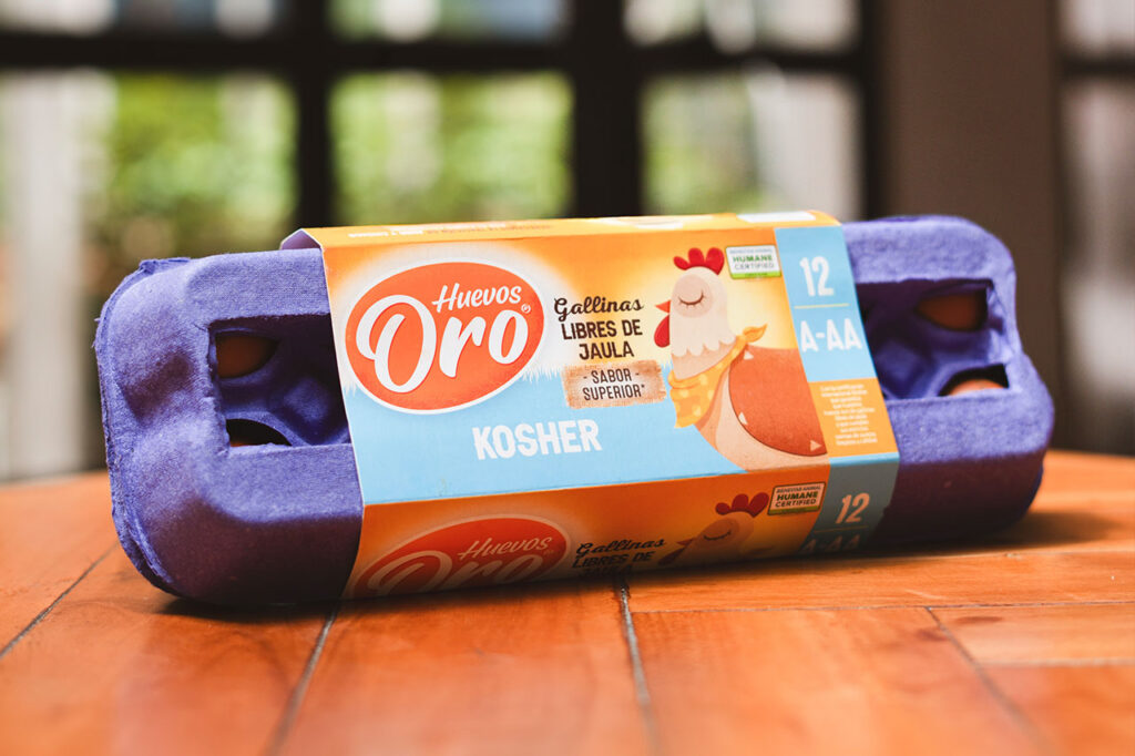Mano sosteniendo un cartón de Huevos Oro Kosher sobre una bandeja de huevos rojos en un entorno de granja.