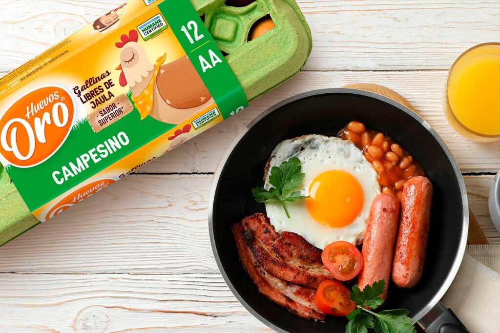 Un desayuno servido en una sartén negra que incluye un huevo frito, dos salchichas, tiras de tocino, frijoles, tomates cherry y perejil. Al lado, se observa un cartón de 12 huevos marca 'Huevos Oro Campesino' de gallinas libres de jaula