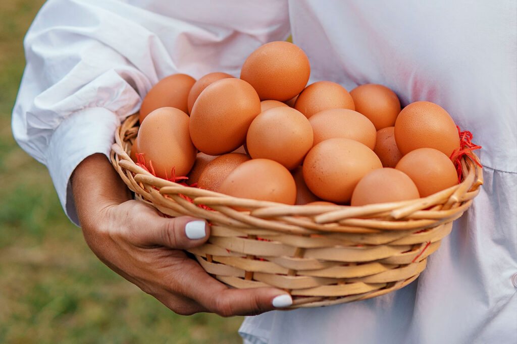 Una persona con blusa blanca sostiene una cesta de mimbre rebosante de huevos marrones lisos y limpios, con un fondo exterior desenfocado