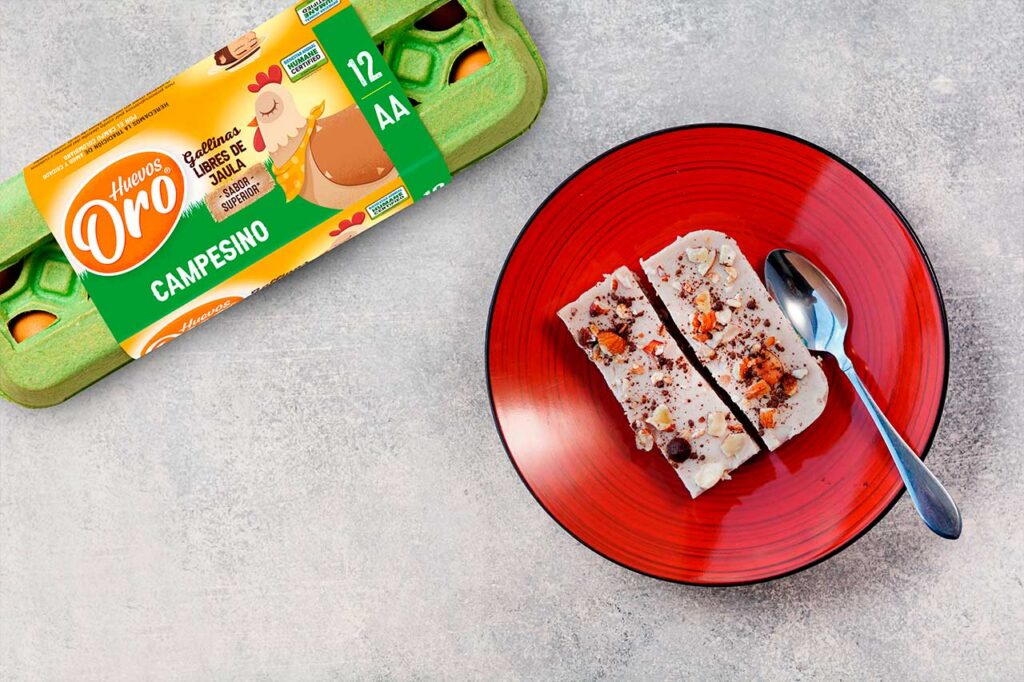 Vista superior de dos rebanadas de postre cremoso espolvoreadas con frutos secos y cacao sobre un plato rojo, junto a una cuchara de metal y un cartón de 12 huevos marca Huevos Oro Campesino.