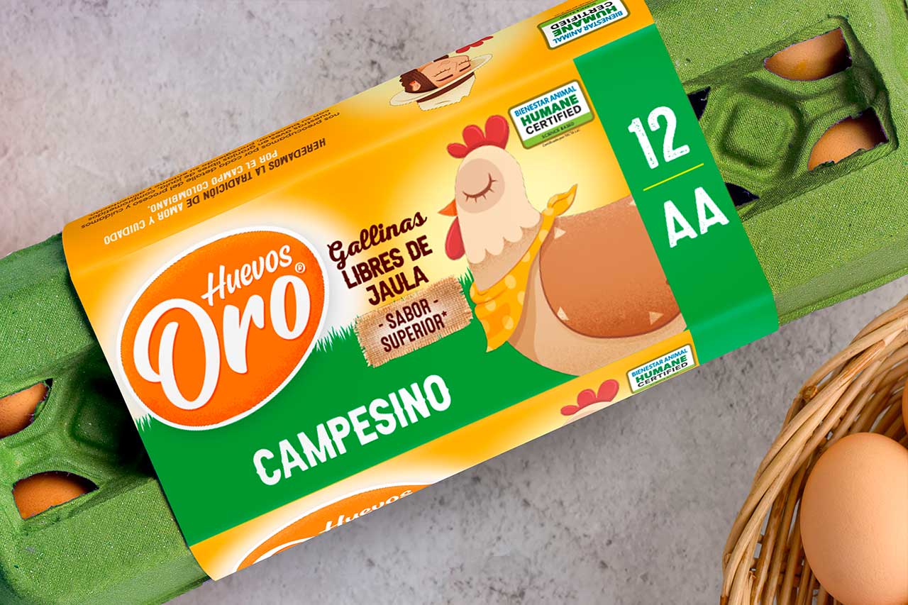 Primer plano de un cartón de 12 huevos marca "Huevos Oro Campesino". El empaque es verde con una etiqueta amarilla que muestra una ilustración de una gallina con pañoleta y sellos de certificación de bienestar animal