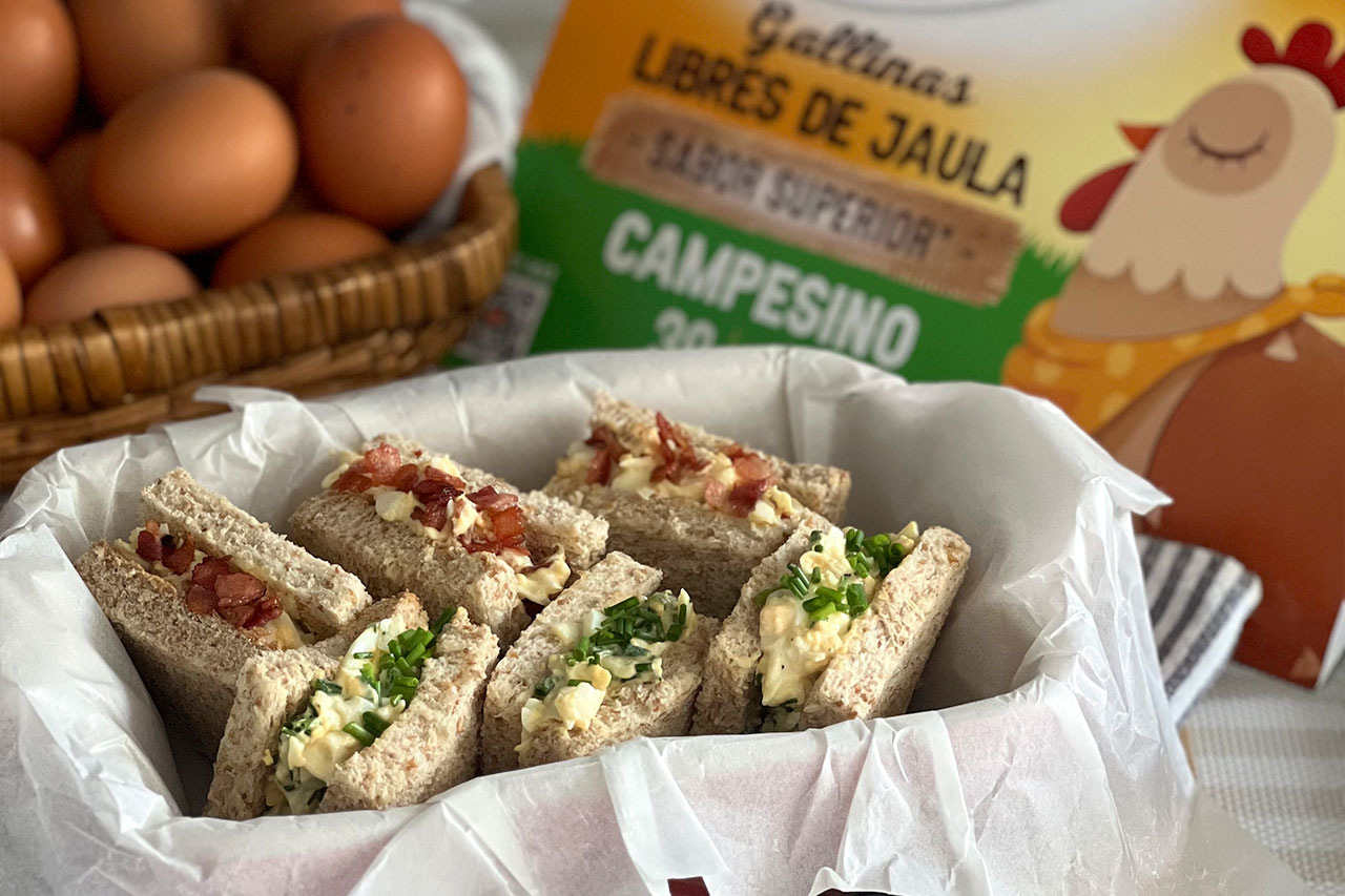 Varios mini sándwiches de pan integral rellenos de ensalada de huevo, organizados en una cesta con papel blanco