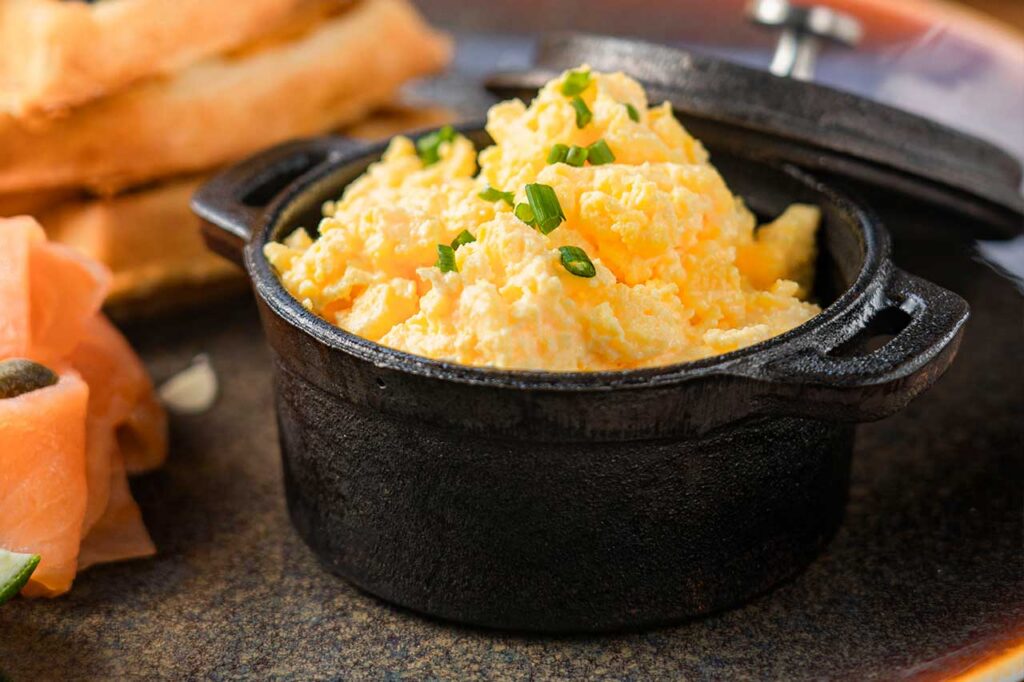 Una porción de huevos revueltos esponjosos servidos en una pequeña cacerola de hierro fundido negra, decorados con cebollín fresco picado.