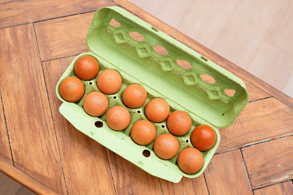 Vista superior de una docena de huevos marrones dentro de un cartón de color verde claro abierto, colocado sobre una mesa de madera rústica